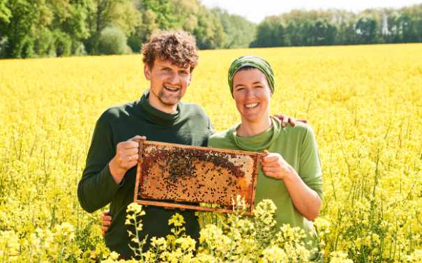 Imkermeisterin Geraldine & J. Friedrich Voigt stehen mit Bienenwabe im Rapsfeld